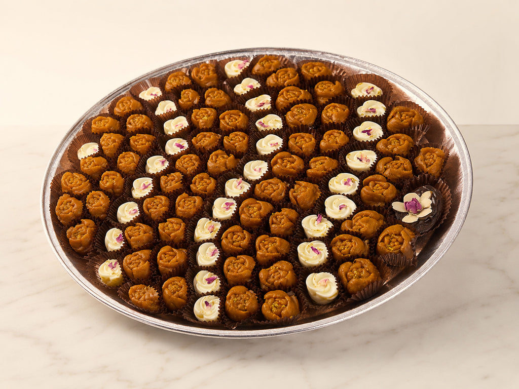 A large round metal tray filled with assorted mini chocolate truffles and molded chocolate pieces, arranged on a light marble surface against a warm off-white background.