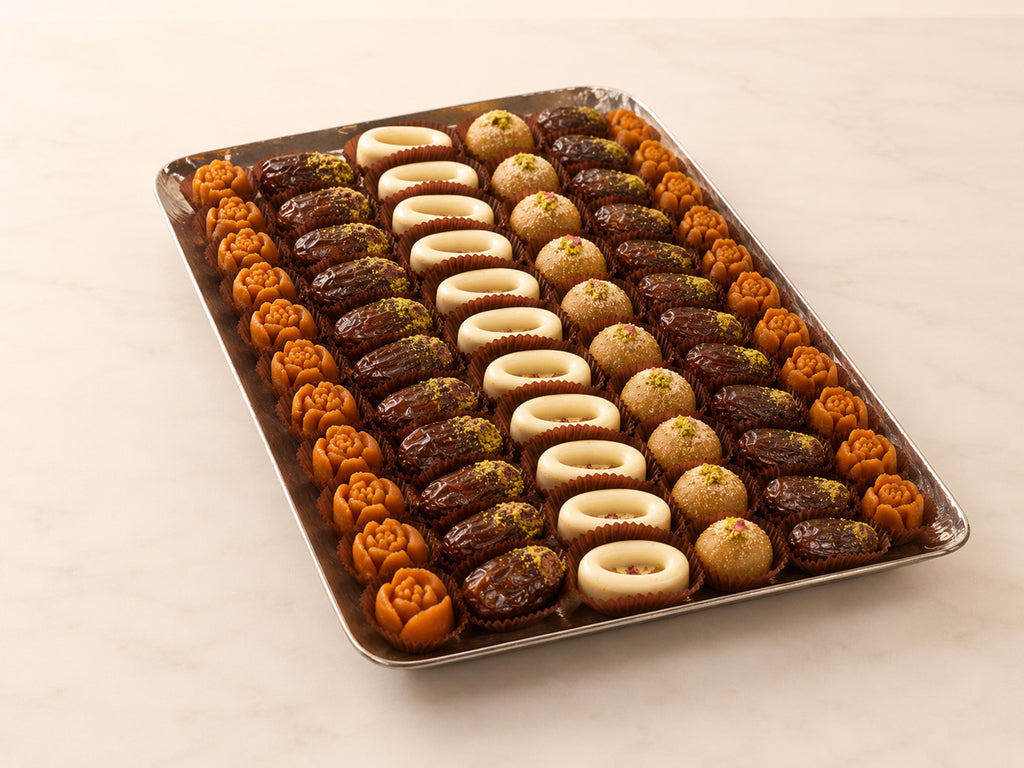 A rectangular metal tray filled with assorted sweet pastries and dates arranged in rows on a light beige marble surface. The pastries include dark chocolate dates, light beige dates, and orange-brown rose-shaped dates.