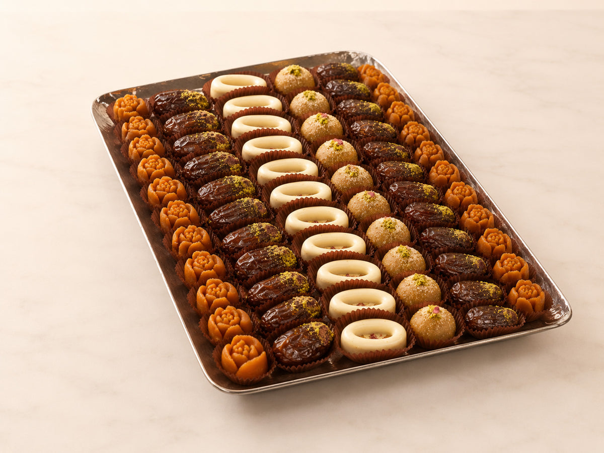A rectangular metal tray filled with assorted sweet pastries and dates arranged in rows on a light beige marble surface. The pastries include dark chocolate dates, light beige dates, and orange-brown rose-shaped dates.